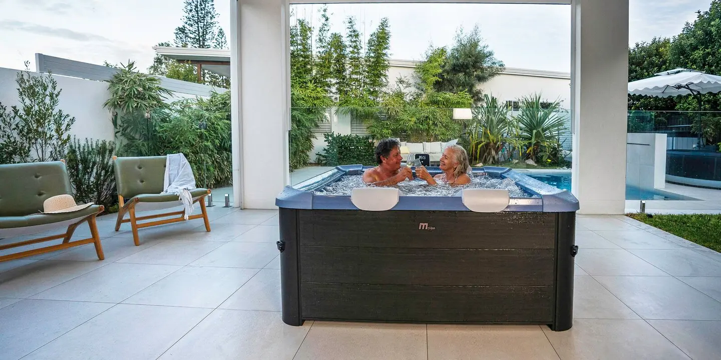 Un couple dans un MSpa Oslo teinte Sapphire, sous une pergola au bord d'une piscine