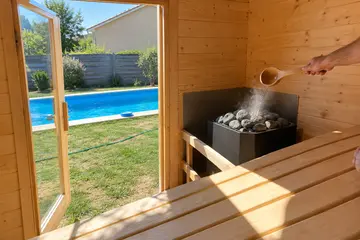 Une louche verse l'eau sur les pierres du poêle, la porte du sauna ouverte sur le bassin