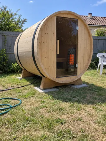Un sauna tonneau au fond du jardin, le poêle rougeoyant derrière la porte vitrée