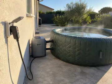 La prise d'un spa gonflable branchée au mur de la maison, l'eau fume derrière