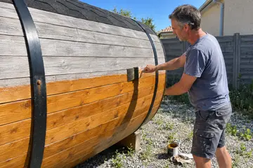 Un propriétaire huile au pinceau un sauna tonneau dont le haut a grisé