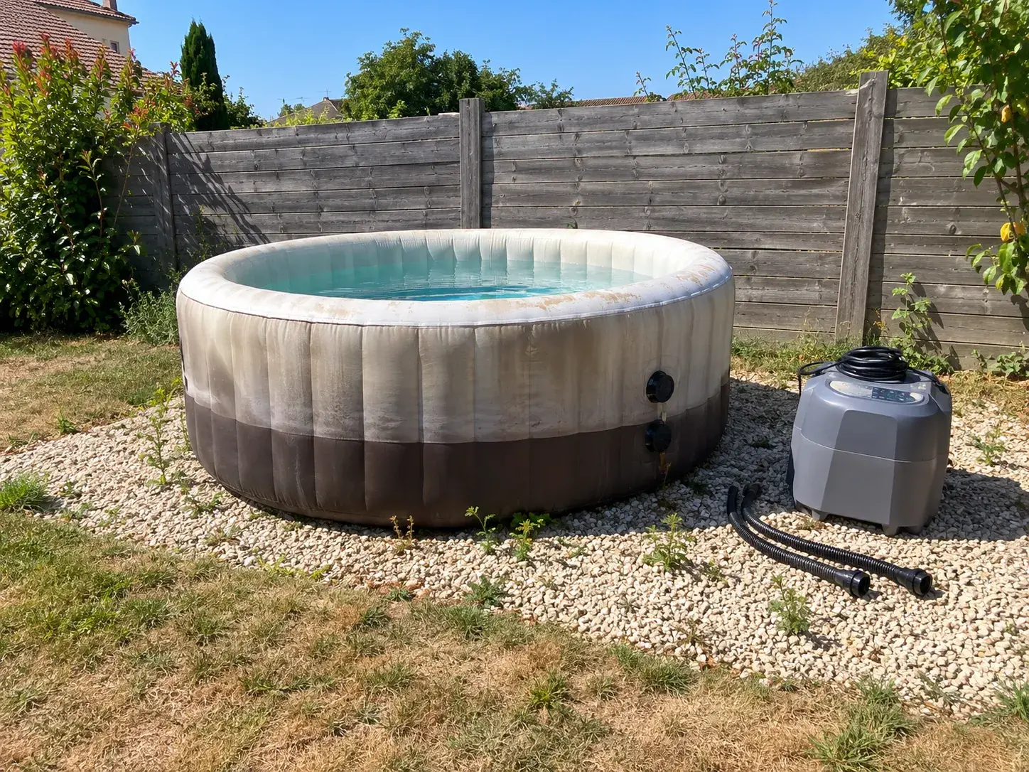 Un spa gonflable délavé par le soleil, son bloc technique débranché posé à côté sur le gravier
