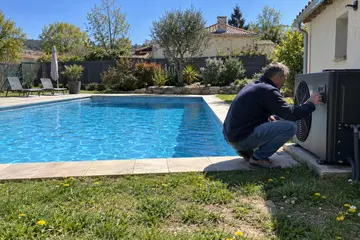 Un propriétaire accroupi règle sa pompe à chaleur au bord de sa piscine enterrée